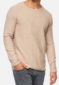 Beiger, texturierter Pullover mit langen Ärmeln und Rundhalsausschnitt. Der Stoff hat ein dezentes Strickmuster, kombiniert mit grauer Hose und Hand in der Tasche.