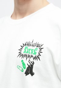 Λευκό βαμβακερό μπλουζάκι με γραφική εκτύπωση που απεικονίζει μαύρα χέρια και πράσινο κείμενο που γράφει "GET LOOSE" και "htf", σε φόντο με σχέδιο εκρήξεως.