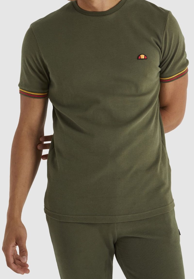 Homme portant une chemise à manches courtes vert olive avec un petit logo sur la poitrine et une bordure rayée sur les manches, associée à un pantalon assorti.
