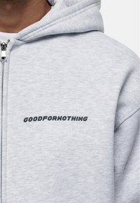 Felpa con zip di colore grigio chiaro realizzata in tessuto morbido, con un audace logo in testo nero "GOODFORNOTHING" sul petto. Dettagli testurizzati sul tessuto.