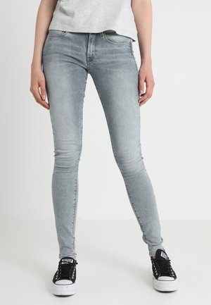 Person trägt hellgraue Skinny-Jeans, ein graues T-Shirt und schwarze schwarze Converse-Sneaker mit niedrigem Schaft und steht vor einem einfarbigen weißen Hintergrund.