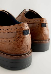 Zapatos de cuero marrón con un acabado texturizado, que cuentan con detalles perforados, una pestaña elástica negra en la parte trasera y una suela negra apilada.