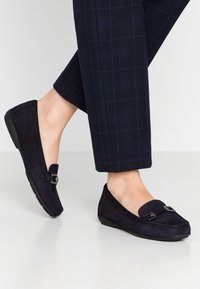 Marineblaue Wildleder-Mokassins mit gesteppten Details, silbernen Hardware-Akzenten und Gummisohlen. Kombiniert mit navyblauen Karottenhosen.