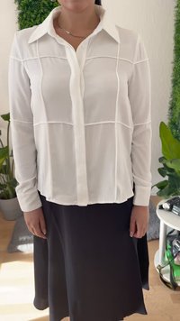 Ženska v beli srajci z dolgimi rokavi in gumbi, ki ima vertikalne in horizontalne šive, v kombinaciji s črno krilo, stoji v zaprtih prostorih ob rastlinah.
