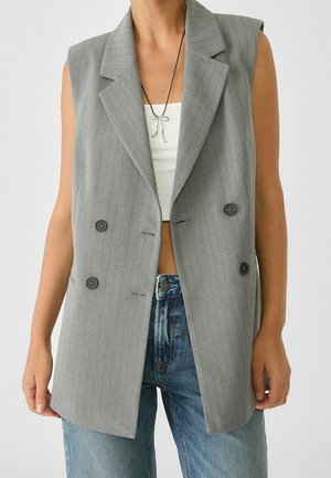 Person trägt einen ärmellosen grauen zweireihigen Blazer über einem weißen Croptop und blauen Jeans, mit silberner schleifenförmiger Halskette an schwarzer Schnur.