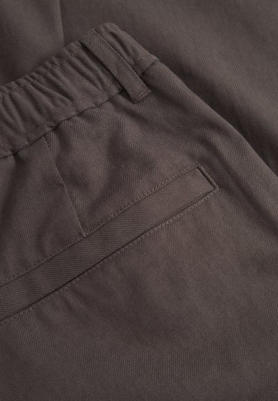 Prim-plan al trousers-ilor maro cu talie elastică, cu bucle pentru curea și cusături vizibile ale buzunarului orizontal.