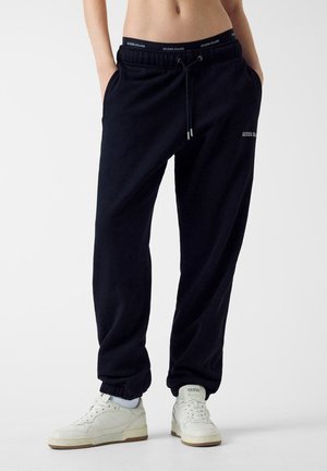 Persona che indossa pantaloni della tuta neri Guess Jeans con coulisse in vita e sneakers bianche Guess, in piedi davanti a uno sfondo semplice.