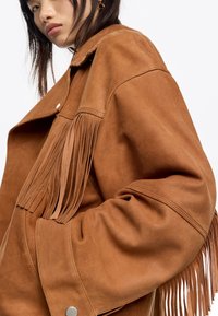 Bruine suedew jacket met franjes langs de schouders en mouwen, met een puntige kraag en zilveren drukknoopsluitingen.