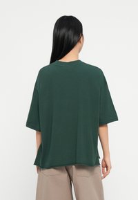 Zöld pamut póló laza fazonban, kerek nyakkal és rövid ujjakkal. Sima textúra, oldalsó hasítékok, bézs nadrággal viselve.