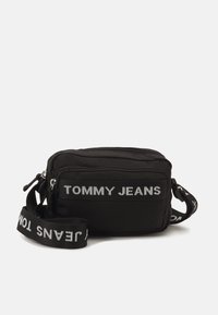 Černá obdélníková crossbody taška s přední kapsou na zip, logo "TOMMY JEANS" na přední straně a nastavitelný ramenní popruh s bílým textem.