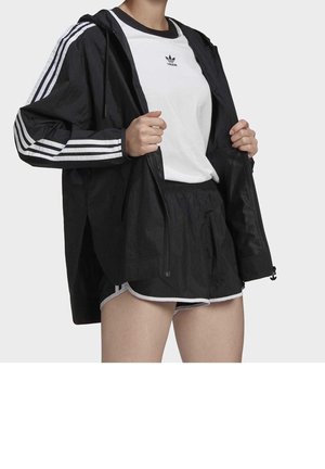 Personne portant une veste noire Adidas avec des bandes blanches, un t-shirt blanc Adidas et un short noir avec des bordures blanches, tenant la veste ouverte.