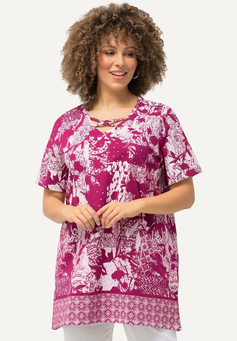 Lila Tunika mit einem auffälligen weißen Blumen- und geometrischen Muster, kurzen Ärmeln und einem V-Ausschnitt mit einem dekorativen Kreuzgurt.