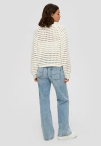 Weißer gestrickter Pullover mit offenem Muster, rundem Halsauschnitt und gerippten Manschetten, kombiniert mit hellblauen, hoch taillierten Jeans und weißen Turnschuhen.