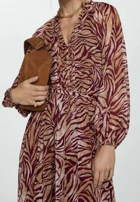 Femme portant une robe à manches longues bordeaux et beige à motifs, tenant un sac à main en daim marron, et portant un collier en chaîne doré et une bague.