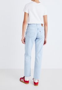 Jeans de mezclilla de color azul claro con corte recto, que cuentan con bolsillos de parche y un parche de logo de cuero en la parte trasera. Combinados con una camiseta blanca y zapatillas rojas.