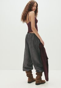 Burgundy stickad topp och lösa, mörkgrå byxor med samlad midja. Bruna slouchy stövlar med remmar. Modell poserar från sidan.