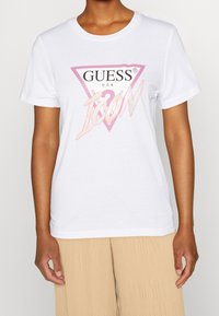 Hvid bomulds t-shirt med et pink trekantet logo, der har "GUESS" og "ICON" i en skinnende skrift. Kortærmet og med rund hals design.