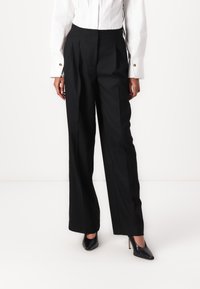 Pantalons larges noirs avec plis à l'avant, taille haute et texture lisse. Associés à des escarpins noirs à bout pointu et une chemise blanche à boutons.
