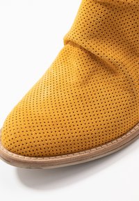 Chaussure en daim jaune avec un design perforé et une semelle beige cousue, gros plan sur la zone des orteils sur fond blanc.