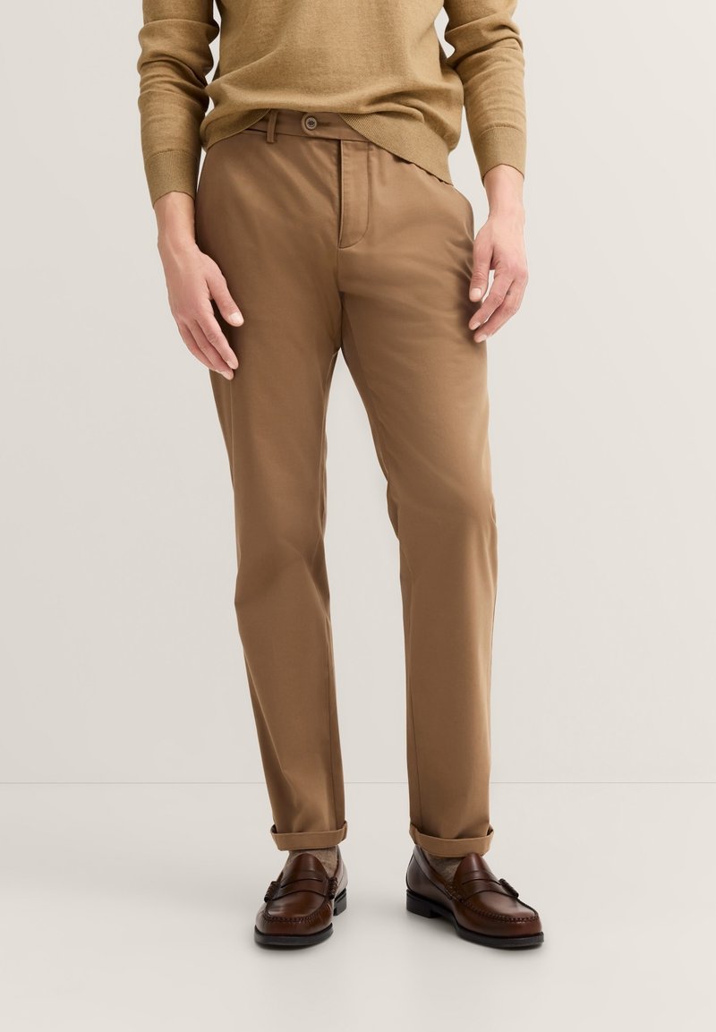 Braune Chinos mit einer glatten Textur, schmaler Passform und einem Knopfverschluss. Kombiniert mit dunkelbraunen Lederschuhen. Nur Frontansicht.