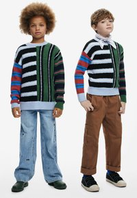 Strickpullover mit kräftigen Streifen in Grün, Schwarz und Weiß; kombiniert mit blauer Jeans und braunen Cargo-Shorts. Weicher Stoff, lockere Passform.