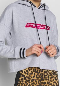 Felpa grigia corta con logo ricamato rosa, lacci neri e polsini a righe. Indossata sopra leggings leopardati.