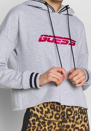 Haut à capuche gris court avec un logo brodé rose, des cordons noirs et des poignets rayés. Porté par-dessus des leggings à imprimé léopard.