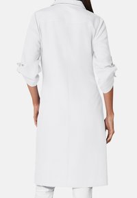 A woman in a white long coat stands facing away, with rolled-up sleeves and brown hair, against a plain white background.