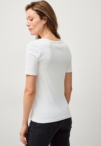Helles, kurzärmeliges T-Shirt aus Baumwolle mit weitem Rundhalsausschnitt und taillierter Passform. Getragen mit dunklen Jeans, die eine glatte Stoffstruktur zeigen.