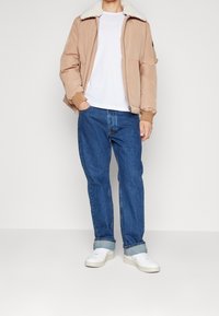 Beige bomberjacka med konstfuskpälskrage, vit t-shirt, blå avslappnade jeans och vita sneakers. Jackan har elastiska manchetter och dragkedja.