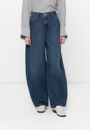 Bredde ben blå denimjeans med høy midje. Materialet ser tykt ut med en slitt overflate. Brukt sammen med en stripete langermet skjorte og lette sko.