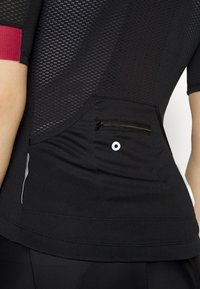 Gros plan d'une personne portant un maillot de cyclisme noir en maille avec une petite poche zippée et un détail réfléchissant blanc sur le bas du dos.