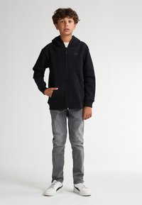 Hoodie zippé noir avec une poche avant et une capuche, porté avec un jean gris et des baskets blanches. Le tissu semble doux et texturé.