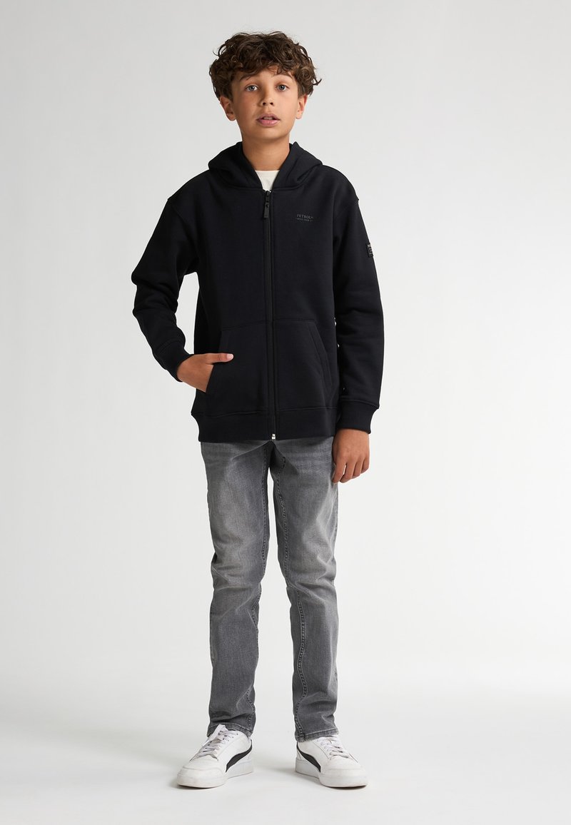 Hoodie zippé noir avec une poche avant et une capuche, porté avec un jean gris et des baskets blanches. Le tissu semble doux et texturé.