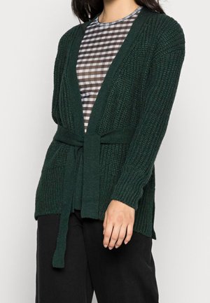 Donna che indossa un cardigan di maglia verde scuro con cintura sopra una maglietta a quadri marrone e bianca e pantaloni neri.