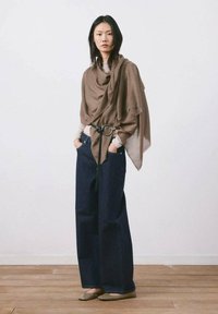 Mujer de pie en el interior, vistiendo un chal marrón drapeado con cinturón, jeans oscuros de pierna ancha, blusa beige y zapatos planos sobre un suelo de madera.