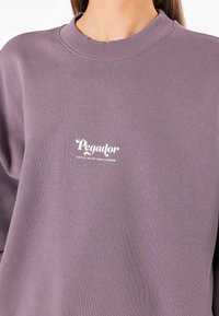 Lilla bomullssweatshirt med ribbet rund hals. Har hvit trykt tekst: "Pegador" og "DETTE ER ALT VI HAR FAMILIE & VENNER."