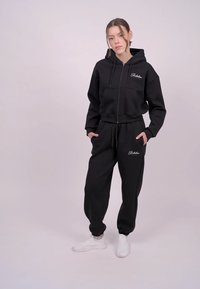 Sort hættetrøje med lynlås, forlommer og logo broderi. Sammen med matchende sorte joggers og hvide sneakers. Blødt, afslappet materiale.
