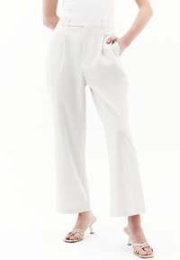 Creme brede broek met voorkruisen, een enkele knoopsluiting en zijzakken, gecombineerd met gouden haksandalen. Gladde stofstructuur.