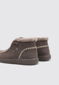 Botas de tobillo de ante marrón con forro de pelo sintético, costuras decorativas, suelas de goma y diseño redondeado con cordones. Los acentos incluyen bordes ondulados.