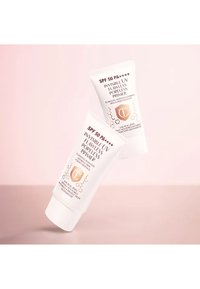 Zwei weiße Primer-Tuben mit goldenen Akzenten, beschriftet mit "Invisible UV Flawless Poreless Primer" und "SPF 50 PA++++", auf einem rosa Hintergrund.