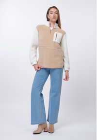 Egy nő bézs és fehér polárpulóvert visel mellzsebbel, világoskék, bőszárú farmert és barna magassarkú bokacipőt.