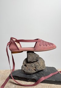 Chaussure plate rose poussiéreuse avec des brides croisées, présentée en équilibre sur des pierres rugueuses empilées sur un fond clair uni.