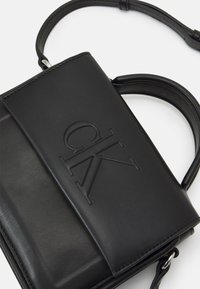 Svart läderaxelväska med en slät yta, med ett präglet logotyp på framsidan, platt design och avtagbar rem.