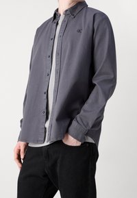 Harmaa kauluksellinen nappipaita, jossa on pieni kirjailtu logo rinnassa, puettuna vaaleamman harmaan paidan päälle ja yhdistetty mustiin housuihin.