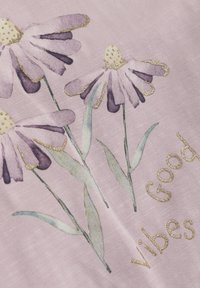 Lys rosa stof har tre lilla blomster med guld accenter og tekst, der siger "Good Vibes" i en skinnende guld skrifttype.