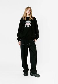Sudadera negra con un gráfico de oso de peluche en gris y rosa. Combinada con pantalones holgados negros y zapatos negros texturizados.
