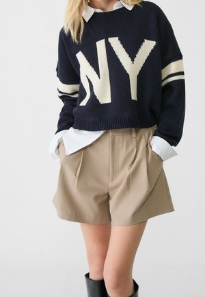 Frau trägt einen marineblauen Pullover mit großen weißen „NY“-Buchstaben, ein weißes Hemd darunter, beige plissierte Shorts und schwarze kniehohe Stiefel.