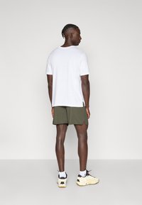 Λευκό μπλουζάκι με κοντά μανίκια και άνετη εφαρμογή, συνδυασμένο με ελιάς πράσινο σορτς από υφή ύφασμα και αθλητικά παπούτσια σε μαύρο και κρεμ χρώμα.