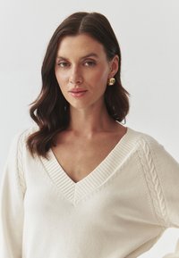 Pulover crem cu decolteu în V și guler cu model în relief, având modele împletite pe mâneci. Material simplu, cu textură.